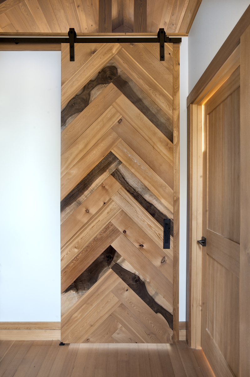 A ‘tall’ flat track door double faced with a herringbone pattern of Walnut, Larch, and Douglas fir is bedroom and hallway wall art for an Oregon couple’s Jewel of a home. Photo by Loren Nelson.